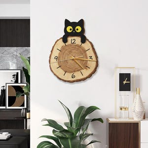Puede incluir: Un reloj de pared de madera con diseño de gato. La esfera del reloj es una sección transversal de un tronco de árbol, con números y manecillas negros. Una silueta de gato negro se encuentra encima del reloj. El reloj mide aproximadamente 25 cm de diámetro.