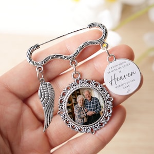 May include: A silver memorial brooch with a safety pin fastening. It features a photo pendant, a wing charm, and a circular charm with the text "I know you'd be with me today if Heaven weren't so far away."