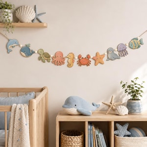 Puede incluir: Una guirnalda decorativa que presenta una serie de recortes de madera de criaturas marinas, incluyendo un delfín, una ballena, una tortuga marina, un pulpo, un caballito de mar, un cangrejo, una estrella de mar, una foca, una medusa y un pez.