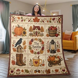May include: A cream-colored quilt featuring a grid of autumn-themed illustrations including a pumpkin, a house, a basket of apples, a crow on a birdhouse, a lantern, boots, a mug with a heart, and a wheelbarrow, with the text The Smiths.