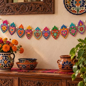 May include: A decorative garland featuring eleven heart-shaped ornaments in vibrant colors like red, blue, green, and yellow, each with intricate floral or eye motifs, strung together on a white cord and displayed horizontally.