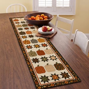 May include: A rectangular quilted table runner featuring a repeating pattern of fabric pumpkins and eight-pointed stars in shades of orange, brown, green, and cream. The runner has a dark floral border and is stitched with a chevron quilting design.