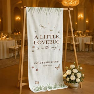 May include: A cream-colored baby shower banner with the text "A Little Lovebug is on the way!" and "Emily's Baby Shower 12.06.2026". The banner features illustrations of butterflies, dragonflies, and flowers. The banner is displayed on a wooden easel.