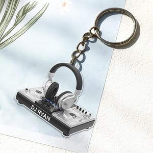 May include: A silver and black DJ keychain featuring a set of headphones and a mixing console. The keychain has the text "DJ RYAN" printed on the console. The keychain is attached to a metal keyring.