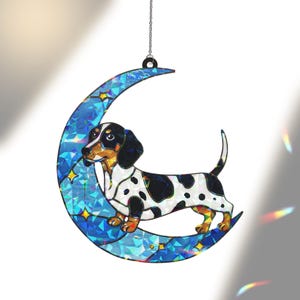 May include: A decorative hanging ornament featuring a dachshund dog standing on a blue crescent moon. The dog has black and white spots, and the moon is decorated with stars. The ornament hangs from a silver chain.