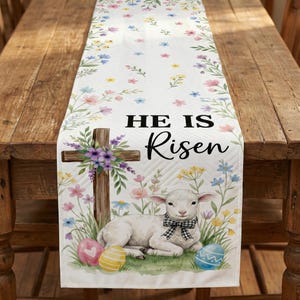 May include: White Easter table runner with floral and leaf patterns. Features a wooden cross, a lamb with a bow tie, and the text "HE IS Risen". Includes colorful Easter eggs and flowers.