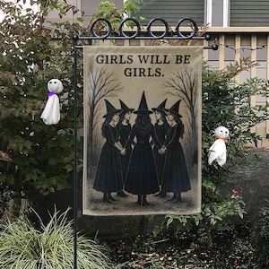 May include: A Halloween garden flag with the text "GIRLS WILL BE GIRLS" and an illustration of five witches in black dresses and hats. The flag is displayed outdoors with decorative ghost figures.