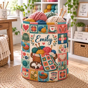 May include: A cylindrical fabric storage basket featuring a patchwork design of yarn-related illustrations, including balls of yarn, knitting needles, a sleeping cat, and a blanket. The centre displays the name Emily in blue cursive script.