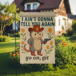 Op de afbeelding: Tuinvlag met een cartoon opossum met een cowboyhoed en een pistool. De vlag toont de tekst "I AIN'T GONNA TELL YOU AGAIN" en "go on, git". De achtergrond is een tuin met bloemen en een huis.