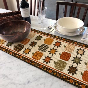 May include: A rectangular quilted table runner featuring a repeating pattern of fabric pumpkins and eight-pointed stars in shades of orange, brown, green, and cream. The runner has a dark floral border and is displayed on a white marble surface.