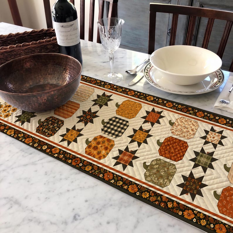 May include: A rectangular quilted table runner featuring a repeating pattern of fabric pumpkins and eight-pointed stars in shades of orange, brown, green, and cream. The runner has a dark floral border and is displayed on a white marble surface.