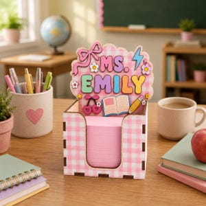 May include: A pink and white gingham patterned notepad holder with the name "MS. EMILY" on top. The holder has a decorative bow, flowers, cherries, a book, and a pencil. A pink heart-shaped container with pens and pencils is in the background.
