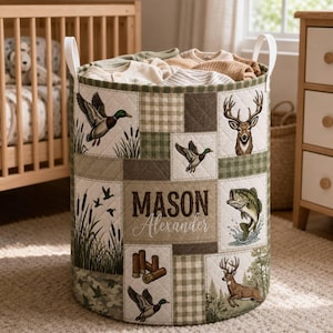 May include: A cylindrical fabric storage basket with white handles, featuring a quilted patchwork design in shades of green, beige, and brown. The exterior displays illustrations of ducks, deer, fish, and shotgun shells, with the name MASON Alexander printed in the center.
