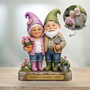 Op de afbeelding: Een grillig tuinkabouterbeeldje met een glimlachend ouder echtpaar. De vrouw draagt een paarse hoed en houdt een pot met roze bloemen vast. De man draagt een groene hoed en houdt een troffel vast. De basis luidt "Grandpa & Grandma's Garden."