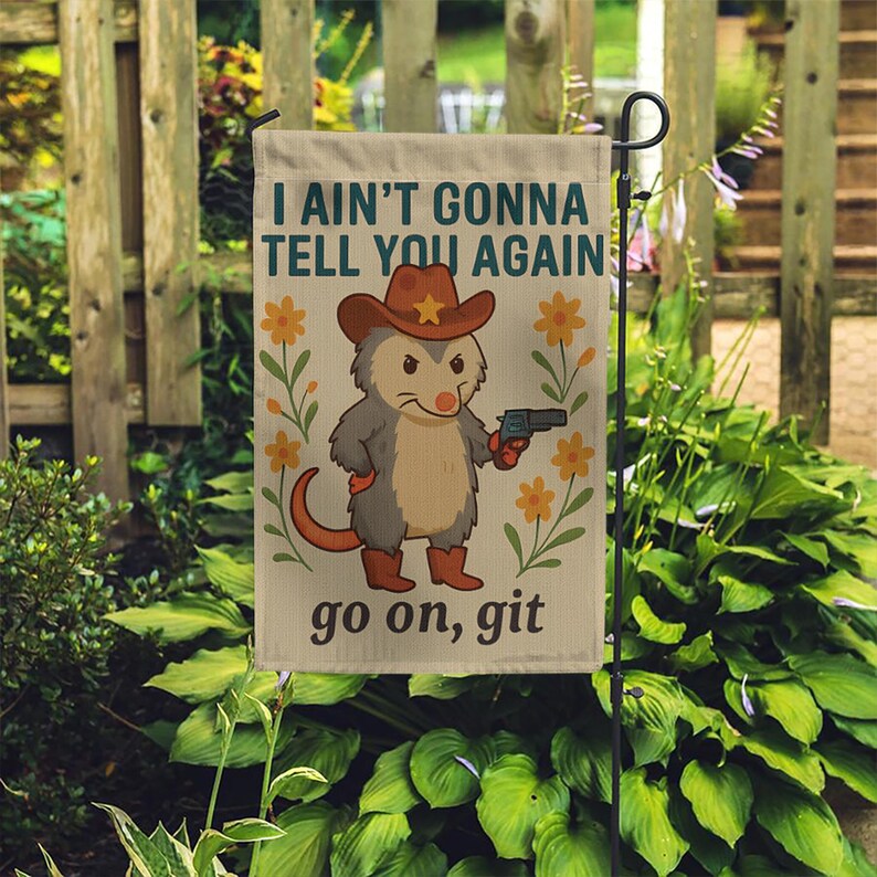 Op de afbeelding: Een tuinvaandel met een cartoon opossum met een cowboyhoed en laarzen, die een pistool vasthoudt. De vlag toont de tekst "I ain't gonna tell you again" en "go on, git", met bloemmotieven.