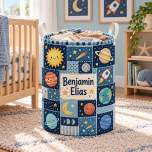 May include: A cylindrical fabric laundry hamper featuring a patchwork design of outer space elements including planets, stars, a sun, a moon, and a rocket ship. The center panel displays the name Benjamin Elias in dark blue text.
