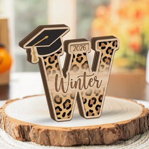 May include: Wooden letter "W" with a graduation cap and leopard print design. The word "Winter" is inscribed on the letter, with the year "2026" above. The letter is displayed on a wooden slice.