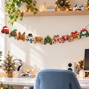 May include: A holiday garland featuring ten decorative gaming controller ornaments strung on a cord. The ornaments include designs of a Santa hat, elf, reindeer, snowman, gingerbread, Christmas tree, peppermint, red sweater, gift, and penguin.