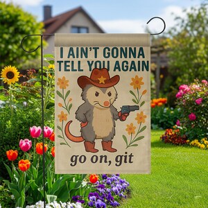 Op de afbeelding: Een beige tuinvaandel met een cartoon opossum met een cowboyhoed en laarzen, die een pistool vasthoudt. De vlag toont de tekst "I AIN'T GONNA TELL YOU AGAIN" en "go on, git", met bloemmotieven.