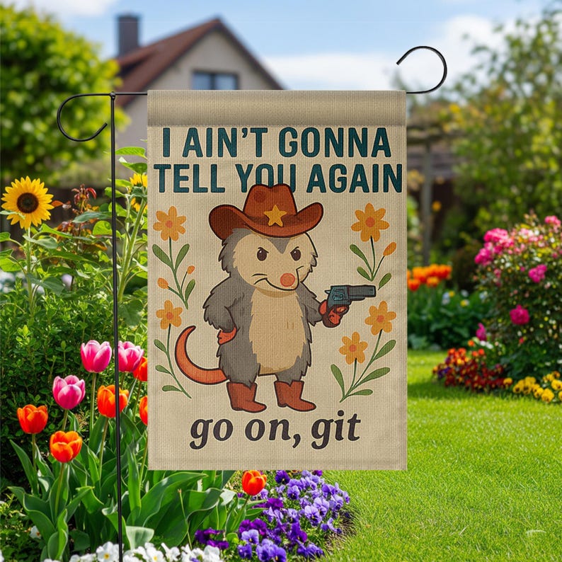 Op de afbeelding: Een beige tuinvaandel met een cartoon opossum met een cowboyhoed en laarzen, die een pistool vasthoudt. De vlag toont de tekst "I AIN'T GONNA TELL YOU AGAIN" en "go on, git", met bloemmotieven.