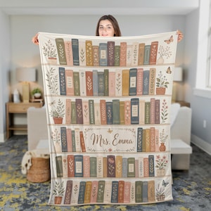 May include: A cream-colored throw blanket featuring a repeating pattern of bookshelves filled with colorful books, potted plants, and butterflies. The center of the blanket displays the text "Mrs. Emma" framed by a delicate floral vine design.