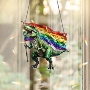 Pode incluir: Um ornamento suspenso com um Tyrannosaurus Rex verde segurando um mastro com uma bandeira arco-íris ondulante. O design tem uma aparência de vitral facetado com cores vibrantes e é suspenso por duas correntes metálicas finas.