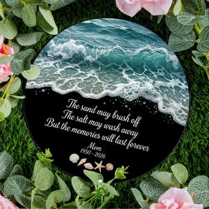 May include: A circular memorial plaque featuring a blue ocean wave crashing onto a black sandy beach. The text reads: The sand may brush off, The salt may wash away, But the memories will last forever, Mom, 1950-2026, with small seashell icons.