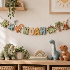 May include: A decorative garland featuring the name NOAH in block letters, interspersed with wooden beads, dinosaur figures, a palm tree, and a volcano. Below, plush dinosaur toys, a wooden rainbow stacker, and a potted plant are displayed.