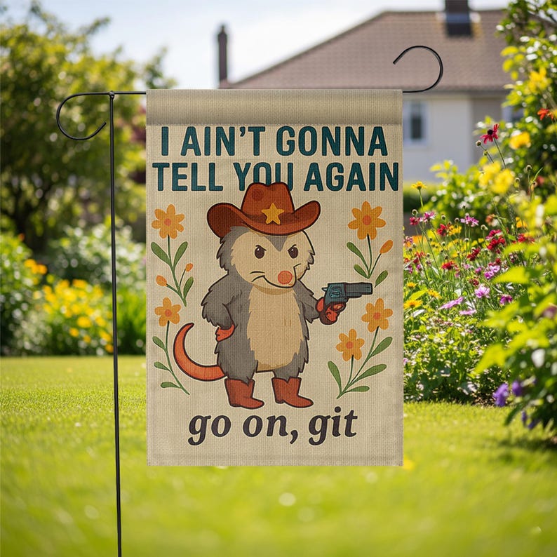 Op de afbeelding: Een tuinvaandel met een cartoon opossum met een cowboyhoed en laarzen, die een pistool vasthoudt. De vlag heeft de tekst "I AIN'T GONNA TELL YOU AGAIN" en "go on, git", met bloemmotieven. De vlag wordt in een tuin getoond.