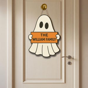 Puede incluir: Un letrero de puerta con temática de Halloween con forma de fantasma, que sostiene un letrero naranja que dice "THE WILLIAM FAMILY". El fantasma es blanco con ojos negros y un contorno negro. Cuelga de un gancho dorado.