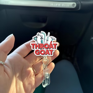 Puede incluir: Un carrete de insignia blanco y rojo con el texto "THROAT GOAT" e ilustraciones de herramientas médicas. El carrete de insignia tiene un soporte de plástico transparente y un clip de metal.