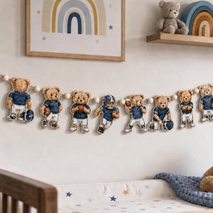 May include: A decorative garland featuring ten individual teddy bear cutouts dressed in blue and white football uniforms, strung together with light-colored wooden beads. Some bears hold footballs, and two are personalized with the name James and number 23.