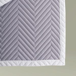 May include: A light gray quilted blanket featuring a repeating chevron pattern stitched across the surface. The blanket is finished with a solid white border that also displays a matching chevron quilted texture along the edges of the fabric.