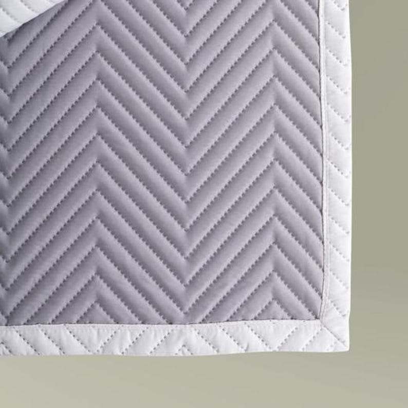May include: A light gray quilted blanket featuring a repeating chevron pattern stitched across the surface. The blanket is finished with a solid white border that also displays a matching chevron quilted texture along the edges of the fabric.