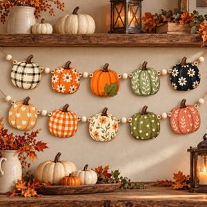 May include: A two-strand garland featuring wooden beads and various decorative pumpkin cutouts. The pumpkins display patterns including black and white plaid, orange with white daisies, solid orange, green with white vines, black with white daisies, hearts, orange gingham, floral vines, and green polka dots.