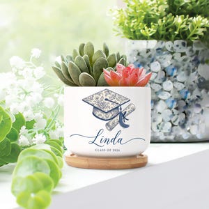 May include: A white ceramic planter with a graduation cap and diploma design in blue, personalized with the name "Linda" and "Class of 2026." The planter holds a succulent arrangement and sits on a bamboo coaster.