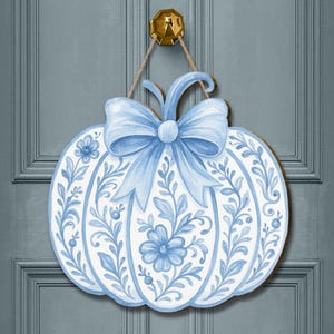 May include: A wooden pumpkin-shaped door hanger featuring a light blue bow and floral design. The pumpkin is painted white with blue floral patterns and a blue bow. A gold door knocker and rope complete the hanging design.