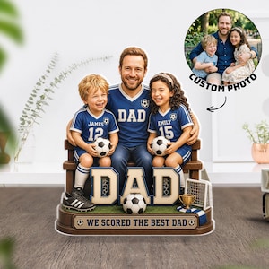 Puede incluir: Una figura 3D personalizada que muestra a un padre y dos niños con camisetas de fútbol azules con el texto DAD, JAMES 12 y EMILY 15. La base incluye un balón, un trofeo, una red de portería y el texto WE SCORED THE BEST DAD.