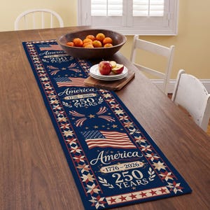 Puede incluir: Un camino de mesa azul marino con un diseño de bandera estadounidense y el texto "America 1776-2026 250 YEARS". El camino de mesa tiene un patrón repetido de estrellas y banderas en los bordes. Un cuenco de madera con naranjas y un plato con manzanas están sobre la mesa.