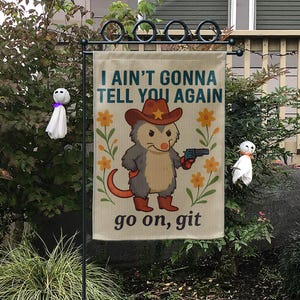 Op de afbeelding: Een tuinvaandel met de tekst "I AIN'T GONNA TELL YOU AGAIN" en "go on, git". De vlag toont een cartoon opossum met een cowboyhoed en laarzen, die een pistool vasthoudt, omringd door oranje bloemen.