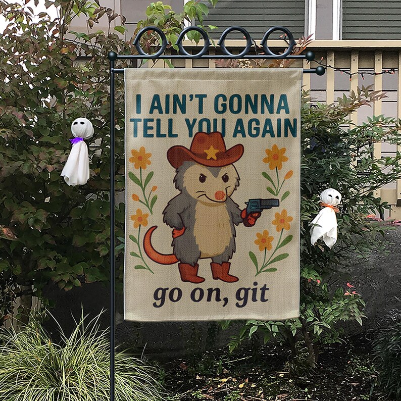 Op de afbeelding: Een tuinvaandel met de tekst "I AIN'T GONNA TELL YOU AGAIN" en "go on, git". De vlag toont een cartoon opossum met een cowboyhoed en laarzen, die een pistool vasthoudt, omringd door oranje bloemen.