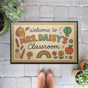 Könnte beinhalten: Eine rechteckige Fußmatte mit dunkelgrünem Rand und cremefarbenem Mittelteil. In der Mitte stehen die Worte "Welcome to MRS. DAISY'S Classroom" in bunten Buchstaben sowie Illustrationen von Schulmaterialien und einem Regenbogen.