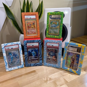 May include: Six framed Pok&eacute;mon trading cards, each with a different design and grade. The frames are painted in various colors, including orange, green, blue, and yellow. The cards are displayed on a wooden surface.