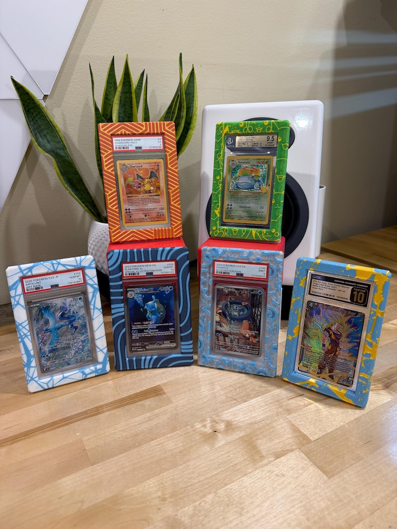 May include: Six framed Pok&eacute;mon trading cards, each with a different design and grade. The frames are painted in various colors, including orange, green, blue, and yellow. The cards are displayed on a wooden surface.