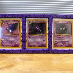 May include: A purple display frame holding three Pok&eacute;mon trading cards: Gengar, Haunter, and Gastly. The frame has a decorative pattern and is made of a solid material. The cards are visible through clear plastic.