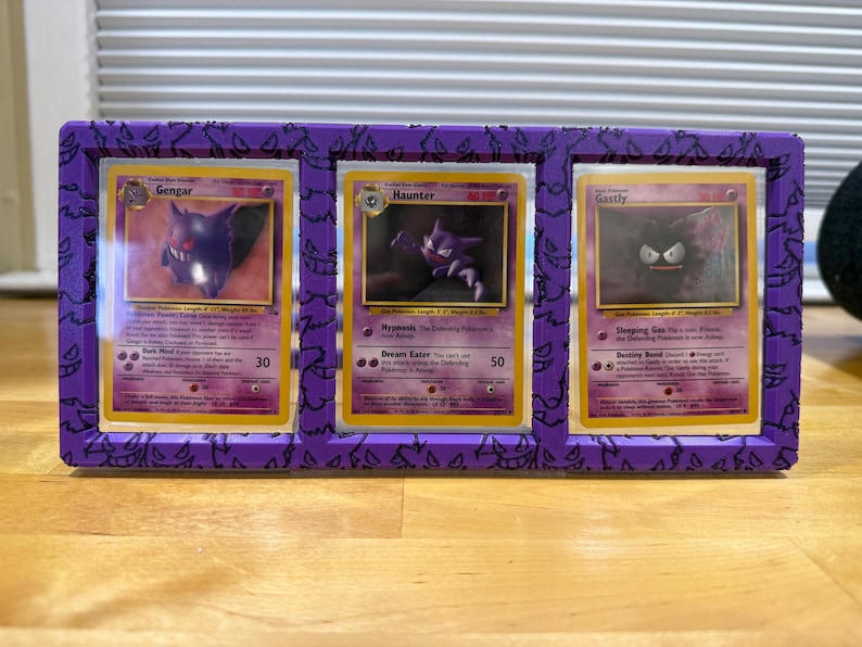 May include: A purple display frame holding three Pok&eacute;mon trading cards: Gengar, Haunter, and Gastly. The frame has a decorative pattern and is made of a solid material. The cards are visible through clear plastic.