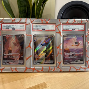May include: A framed display featuring three Pok&eacute;mon trading cards. The cards are encased in protective holders and feature images of Charizard, Charmeleon, and Charmander. The frame has a silver finish with red accents. Text on the cards includes "2023 Pokemon Mew EN" and card details.