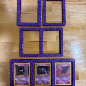Pode incluir: Um conjunto de molduras retangulares roxas com um padr&atilde;o repetitivo preto do personagem Gengar. A cole&ccedil;&atilde;o inclui uma moldura individual, uma moldura individual menor, uma moldura dupla e uma moldura tripla com cartas Gastly, Haunter e Gengar.