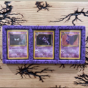 Pode incluir: Uma moldura de exibi&ccedil;&atilde;o retangular roxa com um padr&atilde;o repetitivo do personagem Gengar, contendo tr&ecirc;s cartas colecion&aacute;veis vintage: Gastly, Haunter e Gengar. Cada carta est&aacute; inserida numa capa protetora transparente nas tr&ecirc;s sec&ccedil;&otilde;es.