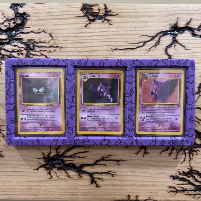 Pode incluir: Uma moldura de exibi&ccedil;&atilde;o retangular roxa com um padr&atilde;o repetitivo do personagem Gengar, contendo tr&ecirc;s cartas colecion&aacute;veis vintage: Gastly, Haunter e Gengar. Cada carta est&aacute; inserida numa capa protetora transparente nas tr&ecirc;s sec&ccedil;&otilde;es.